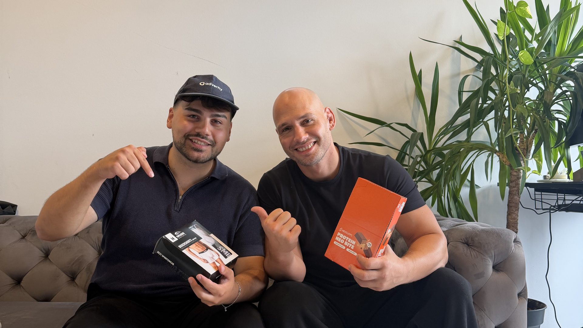 Two of the Neosupps team on the office couch holding Neosupps products, one wearing an Adverfly cap
